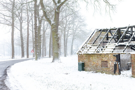 Road passing by burned down barn covered in snow after a blizzard snowstorm with the black wood and construction carcas contrasting against the white hazy winter landscape behind