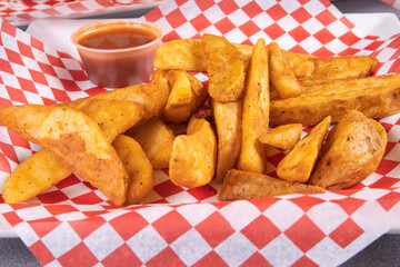 A serving of deep fried coated potato wedges on red checkered paper with red sauce