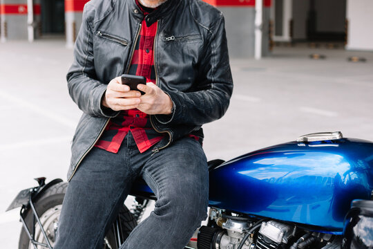 Man in leather jacket checks phone while sitting on blue motorcycle