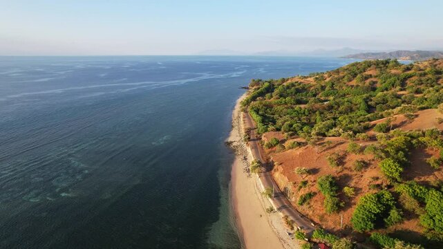 Aerial drone footage of a white sand beach, with asphalt road in the front, and a flat panoramic blue seascape, dry bushes, orange sunset time, in Teluk Gurita, Belu Regency, Timor island, Indonesia