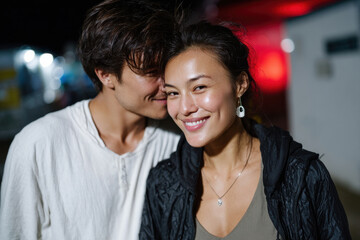 A joyful couple poses closely together outdoors at night, sharing happy smiles as they enjoy each other's company while surrounded by vibrant nightlife colors.