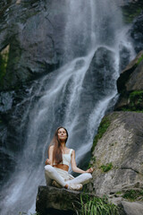 Obraz premium Young woman meditating outdoors near a natural waterfall, wearing white clothes, calm and peaceful expression, surrounded by rocks and greenery in serene nature setting.