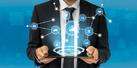 A businessman in a formal suit is holding a tablet that displays a virtual network of technology icons representing connectivity and data analysis in modern business. Tessel