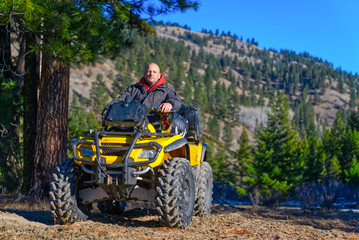 Fototapeta premium Caucasian man with shaved head sitting on a yellow ATV on a sunny day in the forest