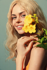 Beautiful woman with long blonde hair holding a yellow flower, exuding joy and freshness against a soft blue background, perfect for summer concepts.