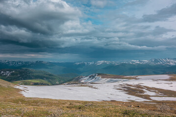 Sunlit snowfield and thawed ground on hill top with view to green hills and big snowy mountain range far away under cloudy sky. Dramatic landscape with sunlight and shadows of clouds high mountains. © Daniil