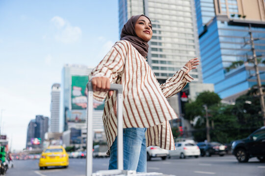 Woman in hijab with luggage, hails a taxi on city street.