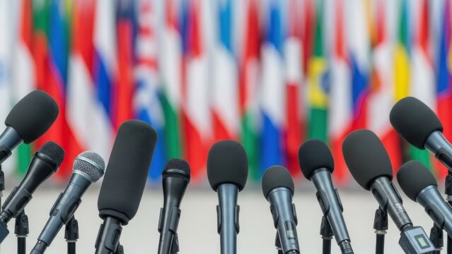 Multiple microphones stand at a podium in front of a colorful array of international flags at a press conference.