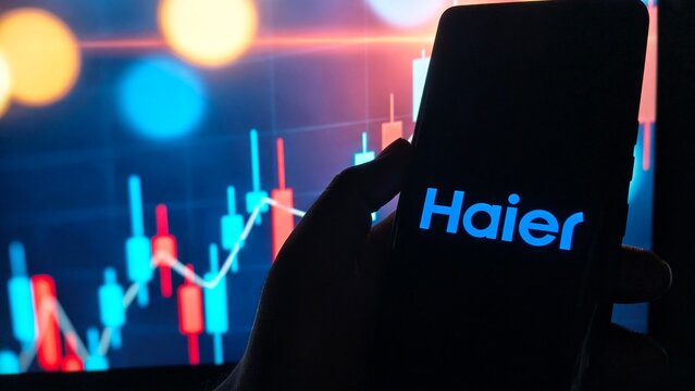Hand holding smartphone with Haier logo in front of stock market graph display
