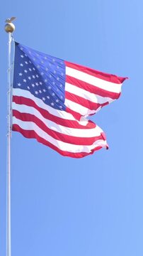 Vertical screen American flag flapping in strong wind, vigorous waves creating dynamic motion, skyline backdrop, civic plaza setting, energetic daylight for parade or ceremony, vibrant red