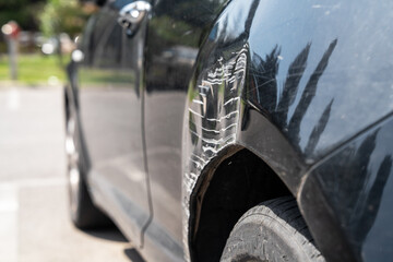 Close-up of a black car with a large dent and deep white scratches on the fender area near the wheel, parked outdoors in a sunlit area with a blurred background