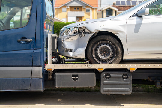 A closer view of the front-left side of a silver car with collision damage and a dented bumper strapped on a tow truck platform in front of a residential neighborhood
