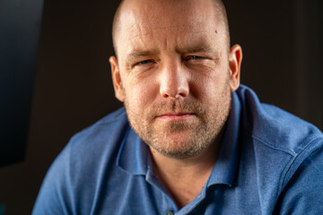 Obraz premium Close up of a Middle-aged white male squinting at the camera wearing a blue casual collared shirt, with plain black background.