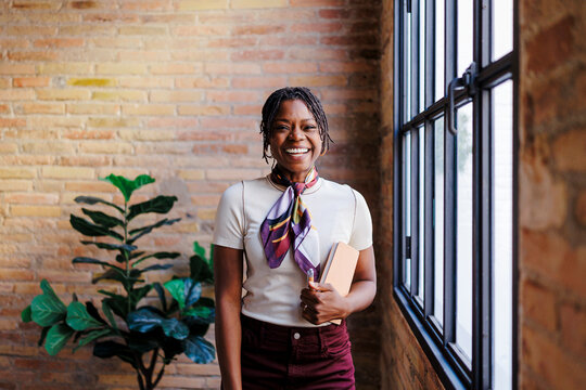 Happy woman holding notebook against brick wall in office