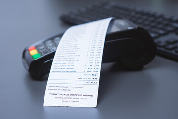 Payment terminal with printed shopping receipt placed on a modern office desk near keyboard. Concept of cashless payment, retail transaction, accounting and consumer expenses.