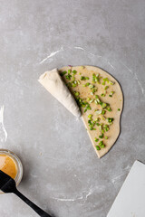 Overhead view of raw chinese scallion pancakes being rolled on grey countertop, top view of homemade scallion pancakes being shaped on countertop, process of making cong you bing