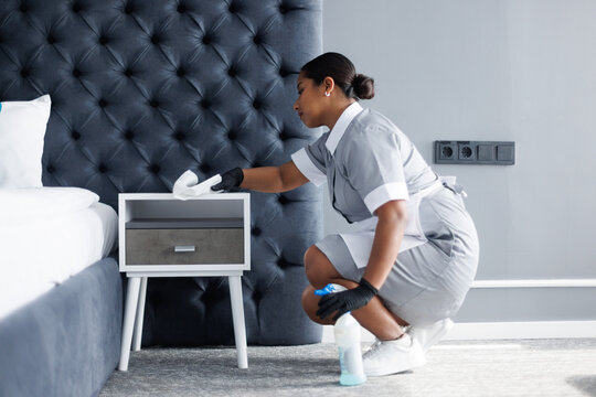Latin maid wiping dust with rag on nightstand in modern hotel room