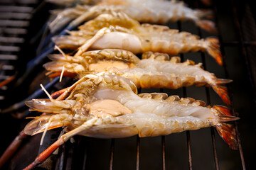 Raw giant river prawns split in half being grilled on a charcoal grill. Close-up shot showing fresh meat and roe, a popular Thai seafood delicacy in preparation.