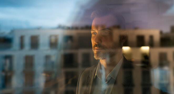 Man reflecting, looking out city window at sunset
