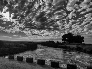 Landscape in monochrome with a low-water bridge, a river, clouds and trees in horizontal format for background use