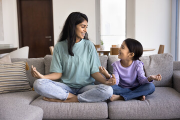 Smiling Indian mom and little 5s daughter sit cross-legged on couch in cozy living room, practicing meditation or mindfulness together. Family bonding through wellness, healthy lifestyle, upbringing
