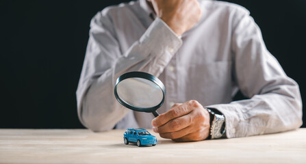 Hand holds magnifying glass above blue car, performing auto inspection, searching vehicle details for insurance claim, concept of finding minute issues requiring careful magnify glass examination