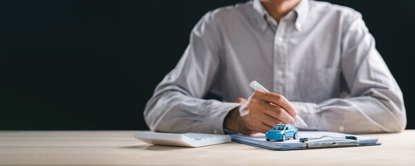 Man signs car purchase contract document, securing auto loan finance insurance, dealership sale lease agreement for new vehicle, calculator and blue model car on desk