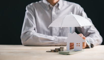 Person shows property protection concept, house model safe under umbrella with coin stacks and money, signifying home insurance and financial security planning