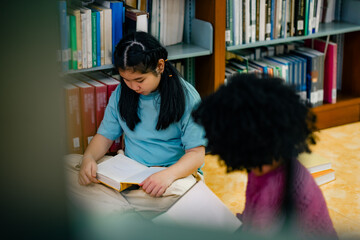 Two children studying quietly inside school library while sharing books on floor. Soft atmosphere reflects concentration friendship learning habits educational growth multicultural classroom concept.