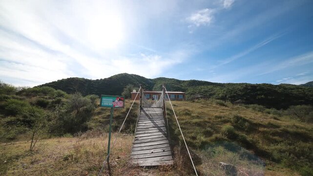 Timelapse de la puesta de sol con un cielo azul y nubes blancas y debajo un puente colgante que va hacia una casa o caba&ntilde;a con fondo de monta&ntilde;as verdes