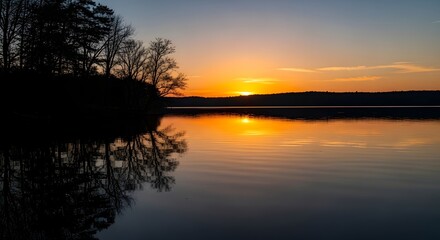 A serene lake at sunset with trees silhouetted against a vibrant orange sky and calm water reflecting the scene.