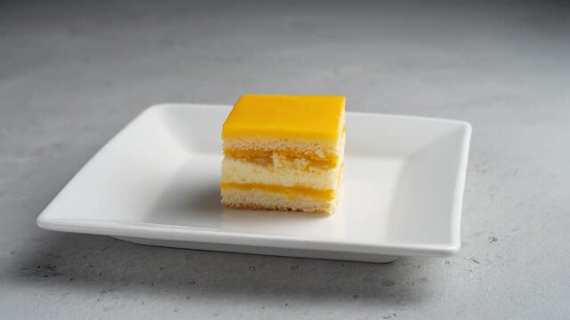 Square slice of layered mango or lemon mousse cake with yellow glaze on a white plate. Smooth 360 orbit rotation for bakery, cafe, and dessert shop commercials.