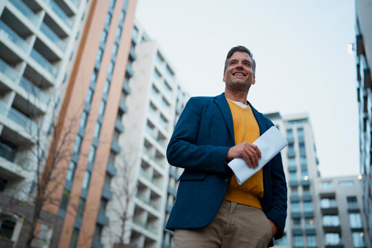 Smiling man holding documents looking at city buildings
