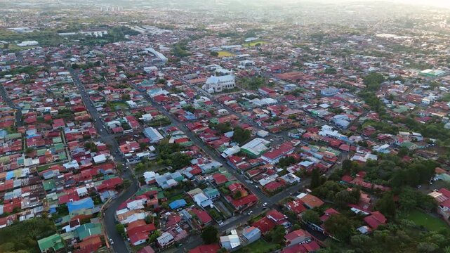 Aerial view flying forward over the residential town of San Rafael, Heredia, Costa Rica, with a prominent white church and red-roofed houses at sunset