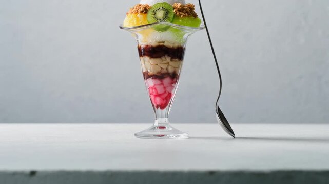 Slow tilt down shot of a tall fruit parfait dessert with kiwi, red beans, and cream in a glass. Elegant sweet treat with a long spoon on a bright background.