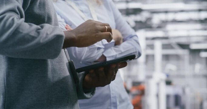 Close Up Hands of Two Safety Supervisors Coordinating Tasks in Bright, Modern Manufacturing Plant. Man Holding Tablet, and Woman Gestures. On Site Communication Efficiency and Adherence to Safety.