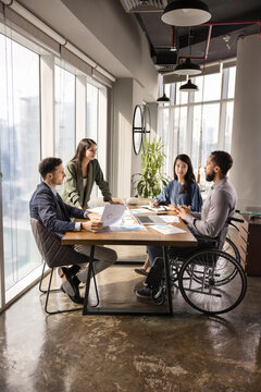 Diverse business team including young African professional in wheelchair discussing project marketing strategy at table, analyzing sales reports together, meeting for brainstorming. Vertical shot
