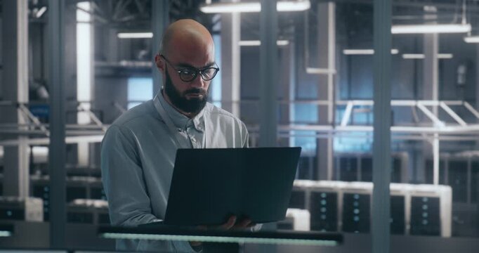 Professional Checking Data Flow on Portable Computer. Young Male IT Specialist Performing System Audit in Aisles of High Tech Facility. Concept of Mobile Technology Use and Data Flow Management.
