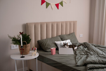 Cat is in interior. Cozy Christmas bedroom interior with green linen bedding, festive decor, and a laptop on the bed. Minimalist holiday home office and winter hygge concept.