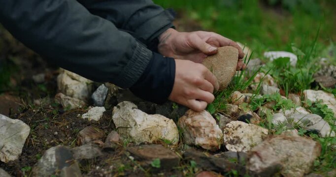 Fingers in dark cuffs slowly envelop smooth stone surface while positioned over loose rock pile with emerging grass shoots, saced ritual preparation in shamanic setting.