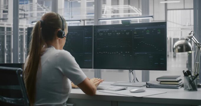 Young Female Professional Manages Large Servers Operations From Modern Workstation. Woman Wears Headset For Active Communication. Technological Innovation, Diverse Workforce and Innovative Workplace.