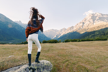 Woman stands on a rock in a wide field looking at jagged mountains, landscape vista in backview...