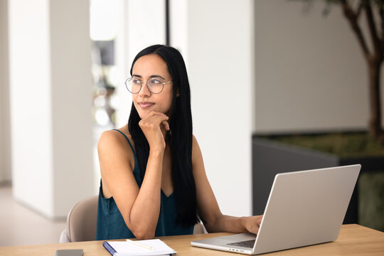 Thoughtful dreamy young Maori freelancer woman in glasses working at computer from home, typing on laptop, looking away, touching chin, thinking on creative solution for online product