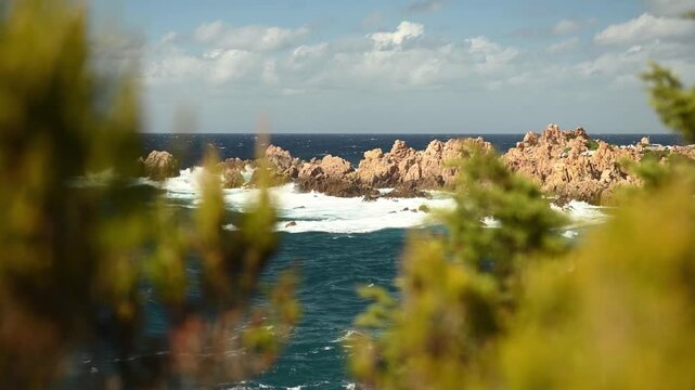 Static video of a rocky coastline in Costa Paradiso, Sardinia, with reddish rock formations and turquoise Mediterranean Sea. Ocean waves break along the shore, while soft foreground foliage adds depth