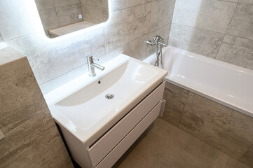 Fototapeta premium minimalist bathroom with a white suspended vanity unit, rectangular sink, built-in bathtub, LED-lit mirror, neutral grey tiles, sleek chrome faucet, clean and modern look.