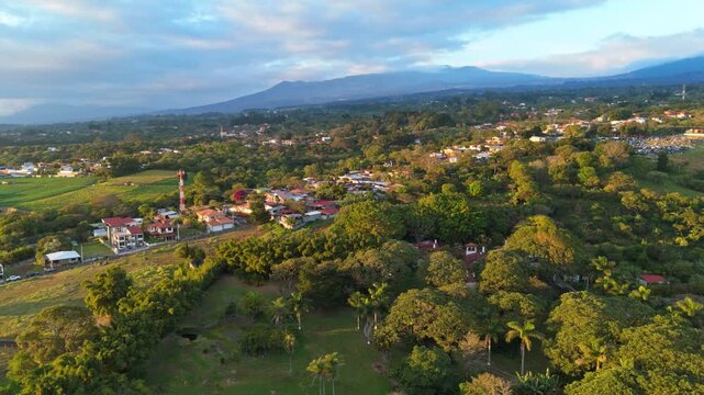 erial flight moving forward over a residential neighborhood in San Rafael de Heredia, Costa Rica, with lush tropical mountains and a glowing orange sunset sky