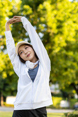 Asian woman wearing wireless headphones, smiling while relaxing in a park after workout and stretching, enjoying music and peaceful outdoor moment of contentment and well-being.