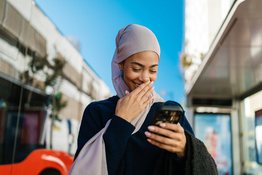 Muslim woman laughing reading smartphone at bus stop