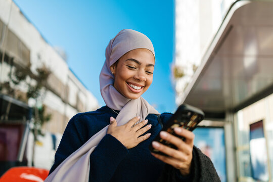Muslim woman happily communicating using smartphone at bus stop