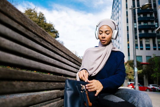 Woman wearing hijab listening music opening bag on bench